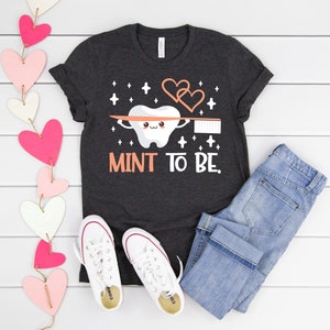 Op de afbeelding: Een donkergrijs T-shirt met een witte cartoon-tand die een tandenborstel vasthoudt en de tekst "MINT TO BE." De tand heeft een schattig gezicht en is omringd door hartjes en sterretjes. Het shirt ligt op een wit houten oppervlak met een paar witte sneakers, een blauwe jeans en een roze-witte hartjeskrans.
