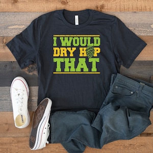 Puede incluir: Una camiseta negra con el texto "I would dry hop that" en letras verdes y amarillas. La camiseta está colocada sobre una superficie de madera con un par de zapatillas blancas y unos vaqueros azules.