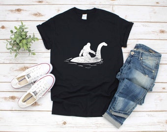 Puede incluir: Una camiseta negra con un gráfico blanco de un Bigfoot montando una criatura similar a Nessie en el agua.