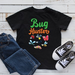 Może przedstawiać: Czarny T-shirt z napisem "Bug Hunter" w zielonych i pomarańczowych literach. Koszulka przedstawia różne kolorowe kreskówkowe owady, w tym biedronki, motyle i pszczoły.