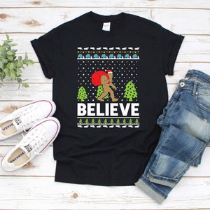 Könnte beinhalten: Schwarzes T-Shirt mit einem weihnachtlichen Design, das einen Bigfoot zeigt, der einen Weihnachtsmann-Hut trägt und einen Sack mit Geschenken trägt. Das Design enthält die Worte "BELIEVE" und ein grünes und weißes Schneeflockenmuster.