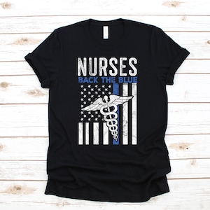 Puede incluir: Camiseta negra con un dise&ntilde;o de bandera estadounidense desgastado que presenta un s&iacute;mbolo de caduceo m&eacute;dico y el texto "NURSES BACK THE BLUE".