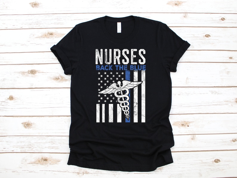 Puede incluir: Camiseta negra con un dise&ntilde;o de bandera estadounidense desgastado que presenta un s&iacute;mbolo de caduceo m&eacute;dico y el texto "NURSES BACK THE BLUE".