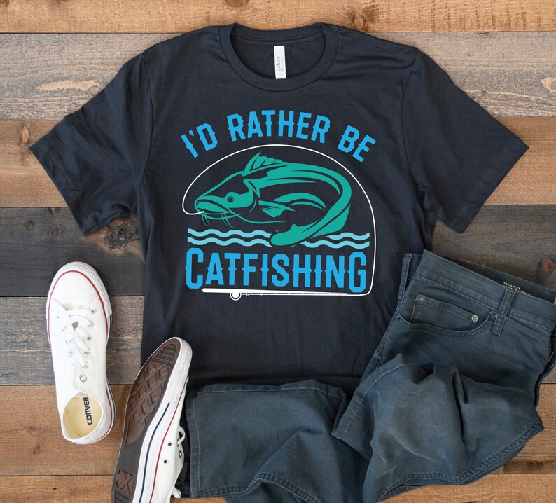 Pode incluir: Uma camiseta preta com um gr&aacute;fico de um peixe-gato e o texto "I'd rather be catfishing". A camiseta est&aacute; colocada sobre uma superf&iacute;cie de madeira com um par de cal&ccedil;as de ganga azuis e t&eacute;nis brancos.