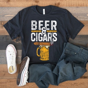 Pode incluir: Uma camiseta preta com o texto "Beer & Cigars" em letras brancas. O design apresenta uma ilustração de desenho animado de uma caneca de cerveja com um charuto no topo.