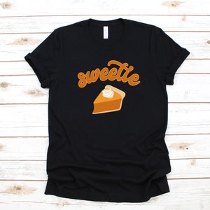 Puede incluir: Camiseta negra con el texto "sweetie" en letra cursiva y una porción de pastel de calabaza con crema batida encima.