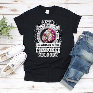 Könnte beinhalten: Schwarzes T-Shirt mit einem Grafikdruck einer amerikanischen Ureinwohnerin mit Kopfbedeckung. Der Text "Never underestimate a woman with Cherokee blood" ist auf dem Shirt aufgedruckt.