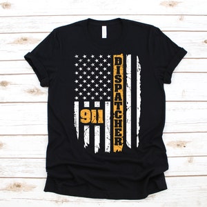 Könnte beinhalten: Schwarzes T-Shirt mit einem abgenutzten amerikanischen Flaggen-Design. Die Flagge hat einen gelben Streifen mit den Worten "911 Dispatcher" vertikal geschrieben.