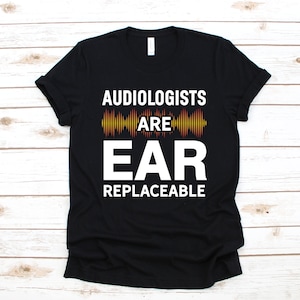 May include: Black t-shirt with white text: "AUDIOLOGISTS ARE EAR REPLACEABLE." The word "EAR" is in a larger font size. Above "EAR" is a graphic of sound waves in yellow, orange, and red. The t-shirt is on a wooden surface.