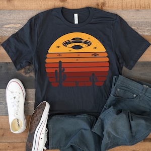 Puede incluir: Una camiseta azul oscuro con un gráfico de puesta de sol retro que presenta un platillo volante y cactus. El gráfico está en tonos de naranja, amarillo y rojo.