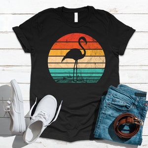 May include: Black t-shirt with a retro sunset graphic featuring a flamingo silhouette. The sunset is in shades of orange, yellow, and blue.