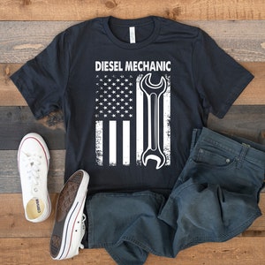 May include: Black t-shirt with a white distressed American flag design and a wrench. The text "Diesel Mechanic" is above the flag.