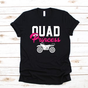 May include: Black t-shirt with the words "QUAD Princess" in white and pink. A white ATV graphic is below the text. The shirt is displayed on a light wood grain surface.