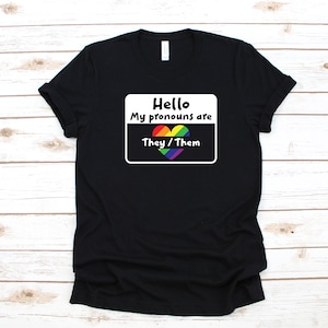 May include: Black t-shirt with a white square that says "Hello My pronouns are They/Them" with a rainbow heart.