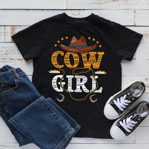 May include: Black t-shirt with a "COW GIRL" graphic featuring a brown cowboy hat, stars, and a rope design. The image includes blue jeans and black canvas sneakers with white laces, all arranged on a white wood-grain surface.