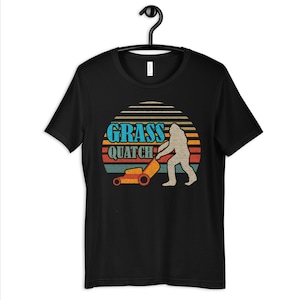 May include: Black t-shirt with a retro sunset graphic featuring a Bigfoot pushing a lawnmower. The text "GRASS QUATCH" is printed in blue and white.