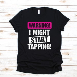 Puede incluir: Camiseta negra con texto blanco que dice "WARNING! I MIGHT START TAPPING!" El texto está en una fuente desgastada. La palabra "WARNING!" está en un cuadro rosa.