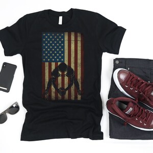 May include: A black t-shirt featuring a vintage American flag design with a silhouette of a football player in a three-point stance.