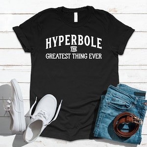 Puede incluir: Camiseta negra con texto blanco que dice "Hyperbole The Greatest Thing Ever". La camiseta está colocada sobre una superficie de madera blanca con un par de zapatillas blancas y unos vaqueros azules con un cinturón marrón.