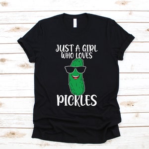 以下が含まれることがあります： 黒いTシャツに、サングラスをかけたピクルスの白いグラフィックと「Just a girl who loves pickles」というテキストがプリントされています。