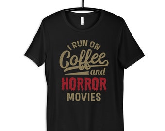 May include: Black t-shirt with a distressed graphic design that says "I run on coffee and horror movies". The text "coffee" is in a brown colour and the text "horror movies" is in a red colour.