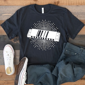 May include: A black t-shirt with a white graphic of a harmonica with a sunburst design. The t-shirt is paired with blue jeans and white sneakers.