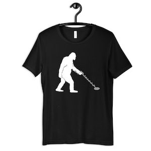 May include: A black t-shirt with a white silhouette of Bigfoot holding a metal detector.