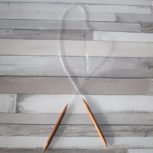 Peut inclure: Une paire d'aiguilles à tricoter en bois avec un câble en plastique transparent. Les aiguilles sont croisées et le câble est bouclé.