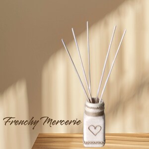 Peut inclure: Un pot en céramique blanche avec un motif en forme de cœur et une bordure en corde brune. Le pot est rempli de bâtonnets diffuseurs blancs. Le pot est posé sur une surface en bois. Le texte "Frenchy Mercerie" est visible dans le coin inférieur gauche de l'image.
