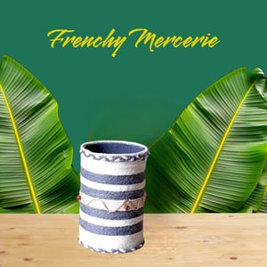 Peut inclure: Un panier de rangement tissé à rayures blanches et grises avec des accents marrons. Le panier est posé sur une surface en bois avec des feuilles de bananier vertes en arrière-plan.