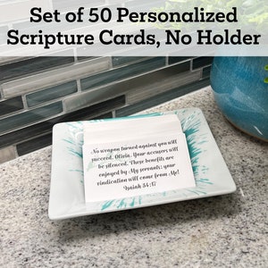 May include: A set of 50 personalized scripture cards in a white rectangular dish with a teal design. The cards are stacked and the top card has a scripture quote from Isaiah 34:17. "No weapon turned against you will succeed, Olivia. Your accusers will be silenced. These benefits are enjoyed by My servants; your vindication will come from Me! Isaiah 34:17"