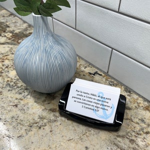 May include: A black card holder with a white card inside. The card features a blue anchor and text in Spanish that reads "Por lo tanto, Nikki, el que est&aacute; unido a Cristo es una nueva persona. Las cosas viejas pasaron y se convirtieron en algo nuevo. 2 Corintios 5:17"