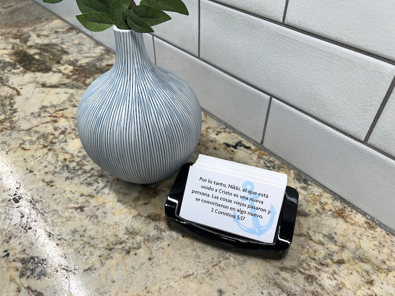 May include: A black card holder with a white card inside. The card features a blue anchor and text in Spanish that reads "Por lo tanto, Nikki, el que est&aacute; unido a Cristo es una nueva persona. Las cosas viejas pasaron y se convirtieron en algo nuevo. 2 Corintios 5:17"