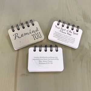 May include: Three small, spiral bound notebooks with inspirational quotes. The first notebook is beige and says "Remind You". The second notebook is white and says "Your Fruit of the Spirit Choice Here" with a quote from Galatians 5:22-23. The third notebook is white and says "Laura, absolutely nothing can separate you from the love of My Son, Jesus Christ. Romans 8:35-39".