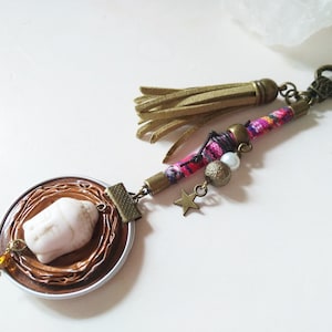 Op de afbeelding: Een bruine en witte Boeddha-hanger met een gouden ster, kralen en een roze en witte kwast bevestigd aan een gouden ketting.