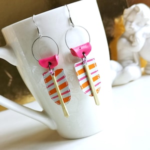 Könnte beinhalten: Handgefertigte Ohrringe mit einem einzigartigen Design. Die Ohrringe haben silberne Reifen, rosa Akzente und rechteckige Anhänger mit orangefarbenen und rosa Streifen. An jedem Anhänger hängt ein goldfarbener Stab. Die Ohrringe werden auf einer weißen Tasse präsentiert.