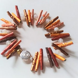 Könnte beinhalten: Eine handgefertigte Halskette mit einem kreisförmigen Design aus metallischen, zylindrischen Perlen in Braun-, Kupfer- und Goldtönen. Die Halskette enthält kleine, klare Perlen sowie silberne und goldene Dekorationselemente.