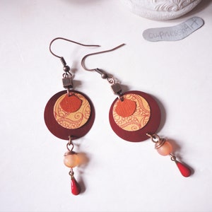Könnte beinhalten: Ein Paar rote und goldene Ohrringe mit einem gemusterten Design. Die Ohrringe haben eine kleine rote Perle und eine größere orange Perle, die daran hängen.