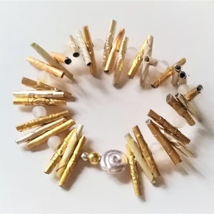 Könnte beinhalten: Ein gold- und silberfarbenes Armband mit einem Muschel-Anhänger. Das Armband besteht aus kleinen, dünnen Metallröhrchen, die mit Gold- und Silberfolie umwickelt sind. Das Armband ist mit weißen Perlen verziert.