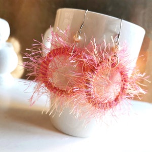 Könnte beinhalten: Ein Paar rosa und weiße, flauschige gehäkelte Ohrringe mit silbernen Haken.