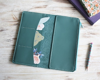 May include: A teal leather-like wallet with a smooth exterior and visible white stitching. The interior features multiple card slots and a zippered compartment, with decorative floral and bear-shaped paper inserts tucked into the pockets.