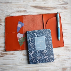 May include: An open orange leather journal with a blue floral patterned notebook and a light blue pen. The journal has a pocket with bear stickers and a green leaf. The notebook has the word "FLOWERY" on the cover.