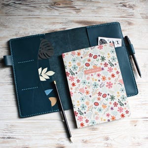May include: A dark teal leather notebook cover with stitched edges, featuring interior pockets and a pen loop. Inside is a notebook with a white cover printed with a colorful floral pattern and the text HIVER BOOKS, plus a black pencil.