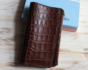 May include: A dark brown wallet featuring a textured crocodile skin pattern. The wallet has a vertical orientation with visible stitching along the edges and a silver-toned metal zipper closure running along the right side of the rectangular body.