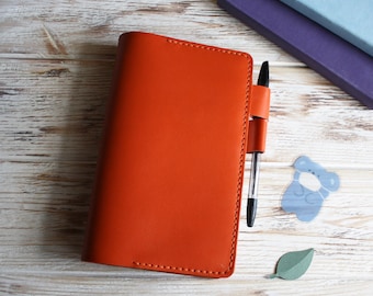 Peut inclure: Un carnet relié en cuir orange avec un porte-stylo et un stylo noir inséré. Le carnet est sur une surface en bois blanc.