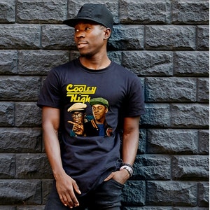 May include: Black t-shirt featuring the text "Cooley High" in yellow and a graphic of two men. The person in the image is wearing a black baseball cap and black jeans. The background is a dark gray brick wall.