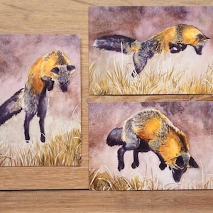 May include: Three watercolor prints of foxes in various poses. The foxes have orange and black fur, set against a backdrop of tall grass and a muted purple sky. The artwork is displayed on a wooden surface.