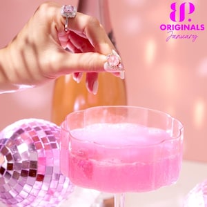 May include: A pink cocktail in a clear glass with a scalloped rim. The cocktail is topped with a pink foam. A hand is holding a pink jeweled ring over the glass, as if to drop it in. The background is pink and features a disco ball and the text "B.P. ORIGINALS January".