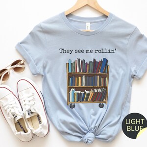May include: Light blue t-shirt featuring the text "They see me rollin'" and an illustration of a rolling bookshelf filled with books.