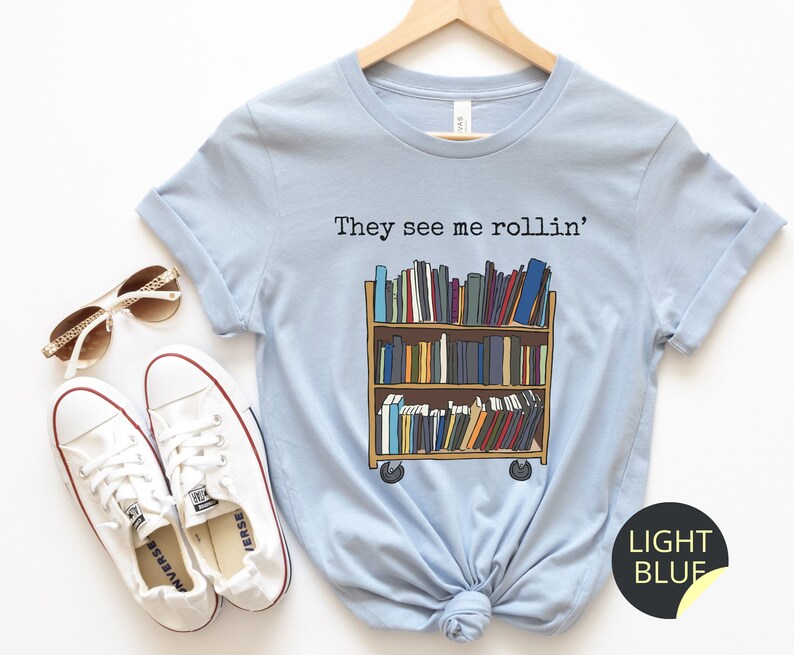 May include: Light blue t-shirt featuring the text "They see me rollin'" and an illustration of a rolling bookshelf filled with books.